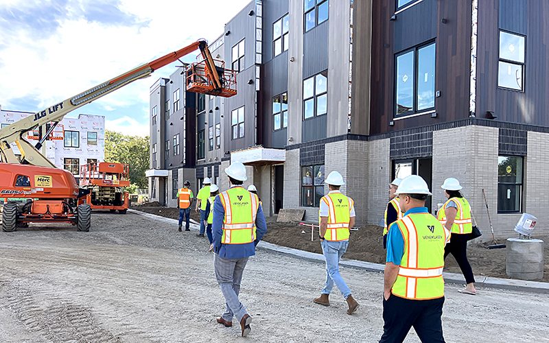 A group of people in hard hats and high-visibility vests walk through an active construction site, passing a multi-story building under development and a boom lift in use.