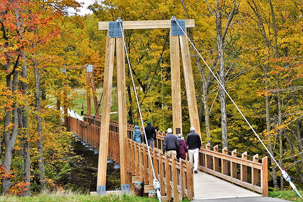 Five older people cross a suspension bridge through a forest in autumn.