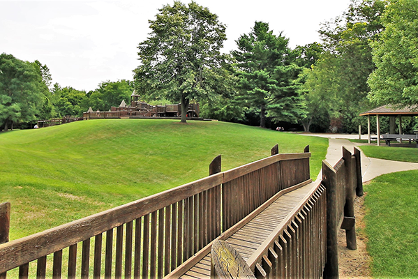 A foot bridge leads to a playground and a covered pavilion in a partially wooded area.
