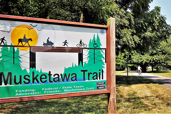 The Musketawa Trail marker; A person bikes along a trail in the background