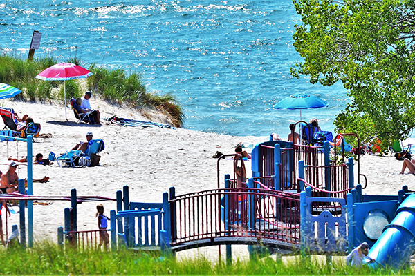 Children play on a large playground at the beach; Families sit in the sand and sun behind them