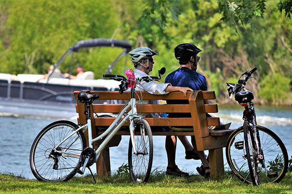 A couple rests on a bench with their bikes nearby; A man operates a pontoon boat in the water behind them.