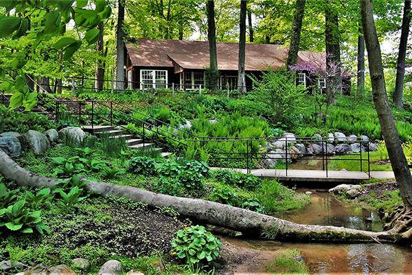 The picnic building overlooks a small creek and footbridge; It is surrounded by spring foliage and plants.
